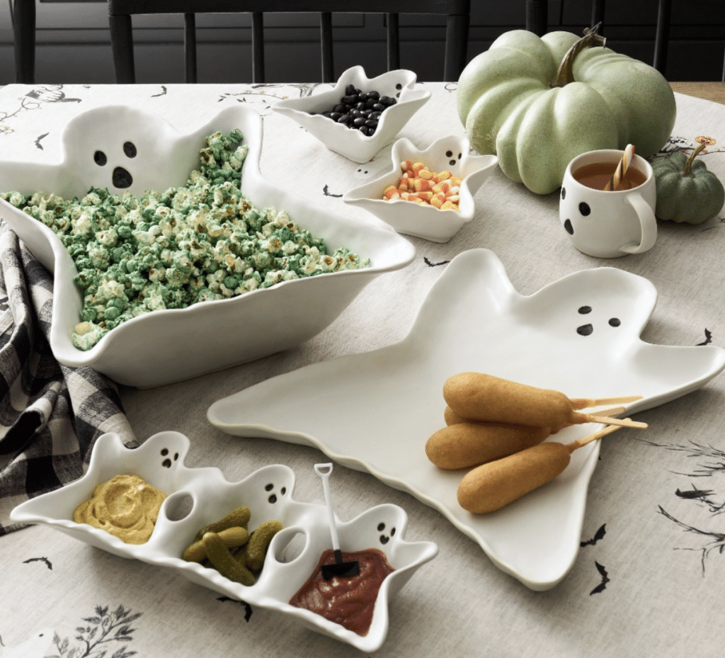 A Halloween-themed table setting delights with ghost-shaped platters featuring halloween treats for kids like green popcorn, mustard, corn dogs, and pickles. There's also a black broomstick stirrer in a condiment, candy corn, a pumpkin mug, and a green decorative pumpkin.